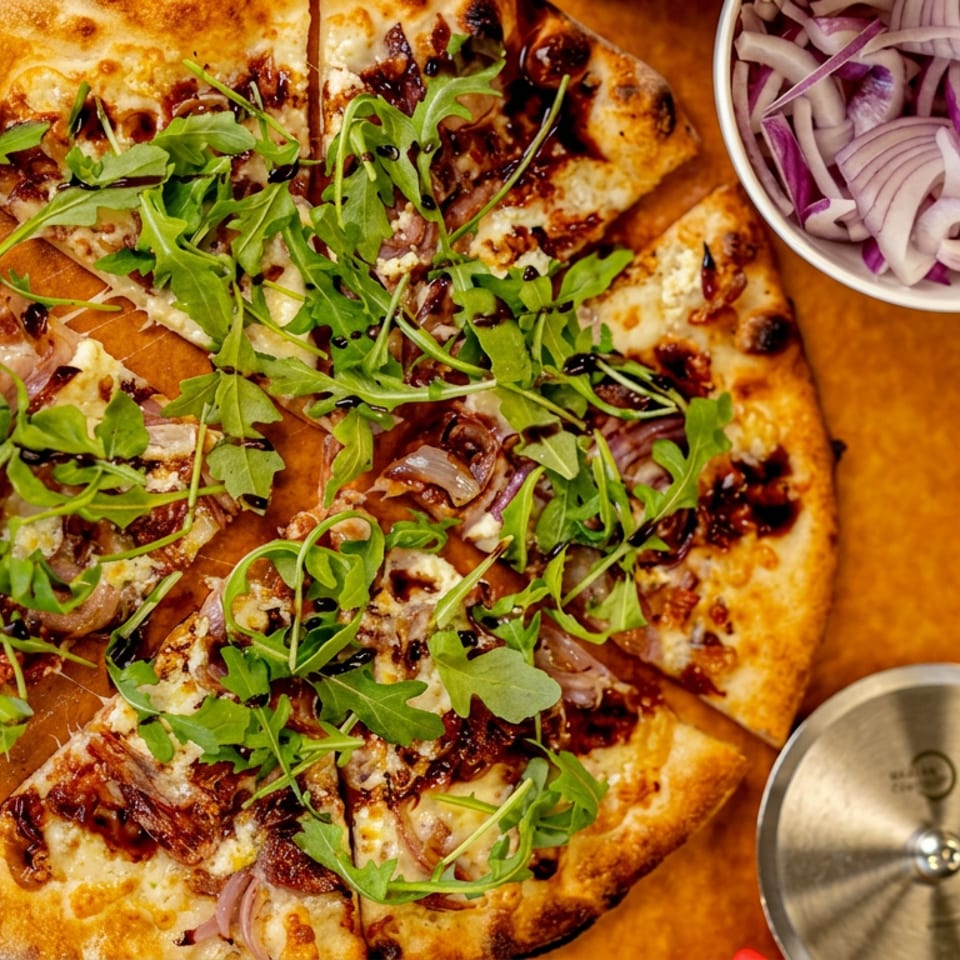 Overhead view of sliced flatbread pizza topped with arugula, caramelized onions, and balsamic glaze, served with pickled onions.