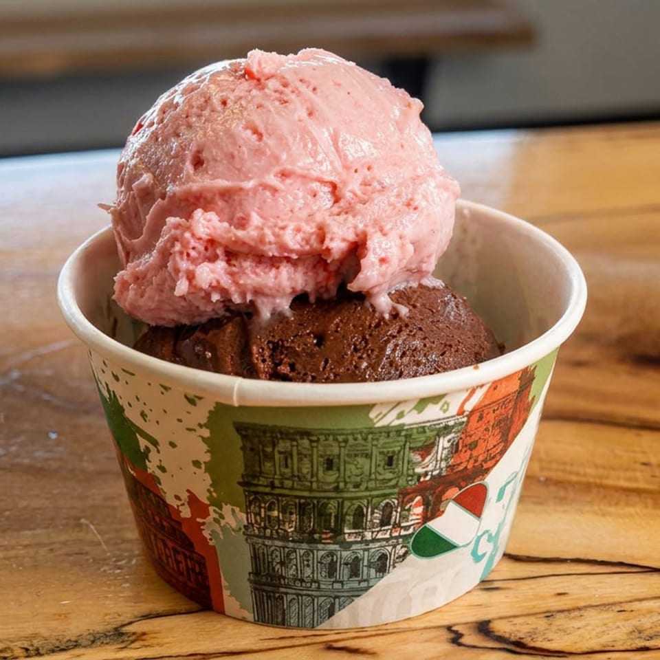 Two scoops of pink and chocolate gelato in a colorful paper cup on a wooden table.