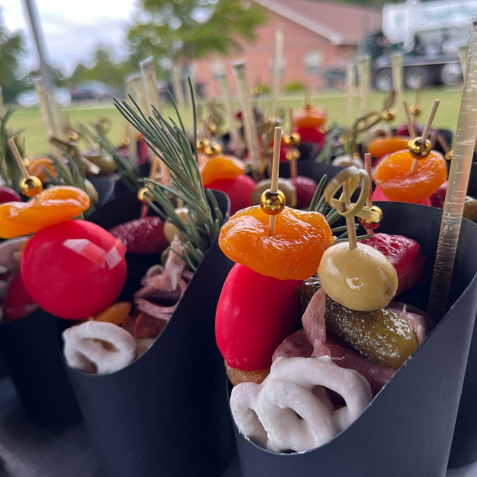 Colorful appetizer skewers with tomatoes, citrus, olives, pickles, and herbs arranged in black containers outdoors.